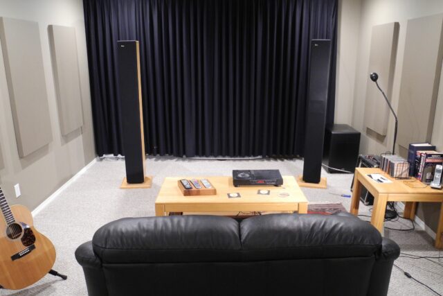 view behind couch in home theatre with table and speakers in front and grey acoustic panels on side walls.