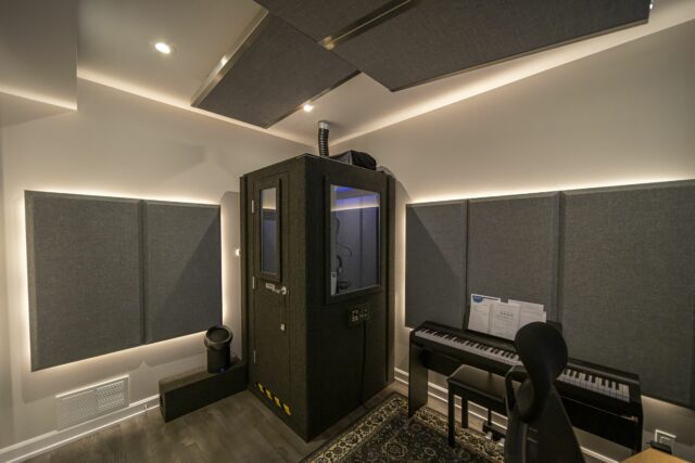 Vocal booth in corner of studio room with piano on the right and grey acoustic panels on walls and ceiling