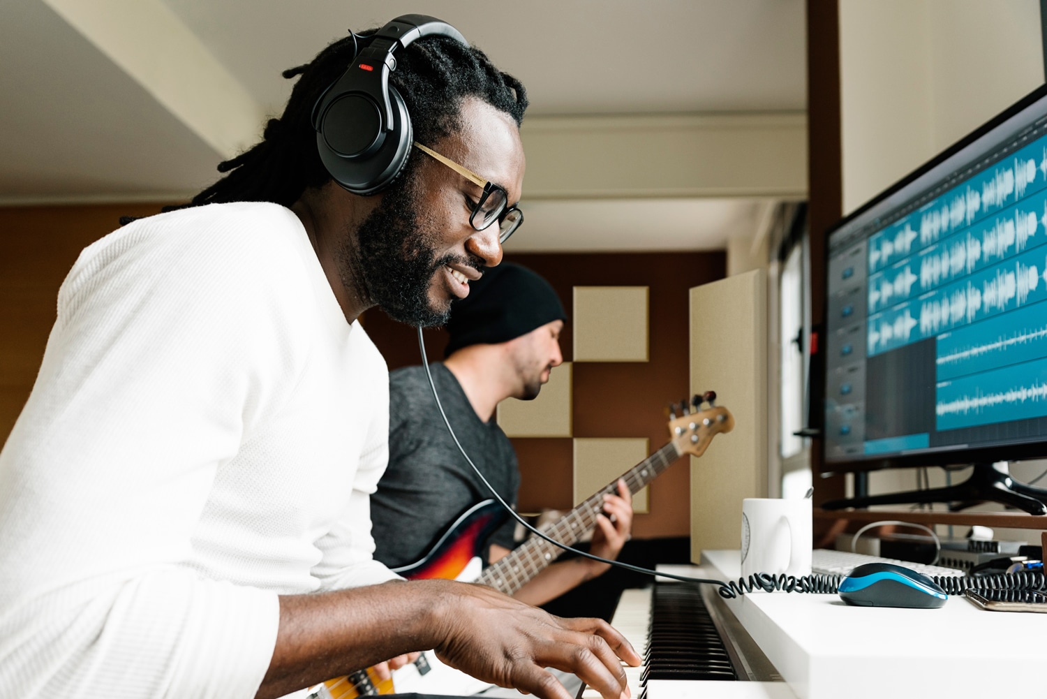 Two men playing instruments in a recording studio with beige acoustic panels on back wall.