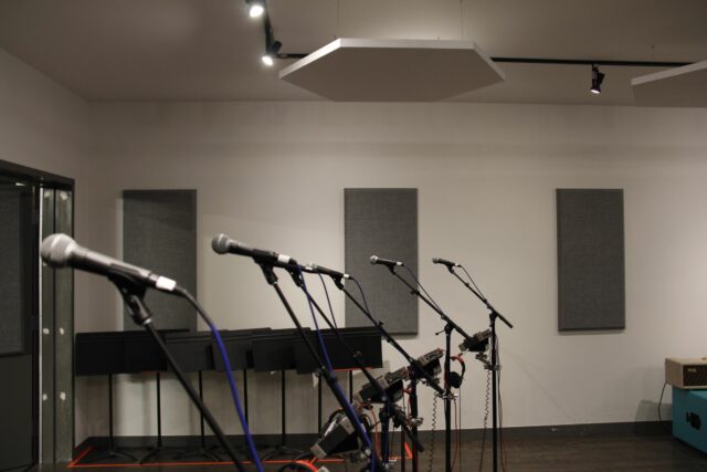 Multiple microphones ready for recording with grey acoustic panels on back wall and white hexagonal acoustic panels on ceiling