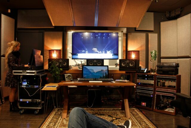 First person view of man on couch looking onto recording studio mixing station with woman standing on the left and acoustic panels hanging on ceiling above