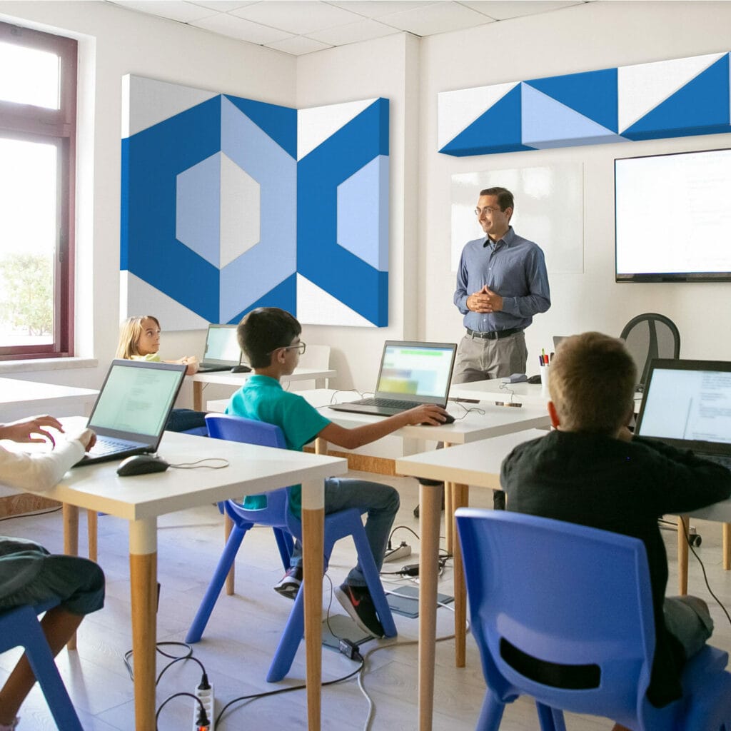 Male teacher speaking to class of elementary students with white and blue acoustic panels on wall.