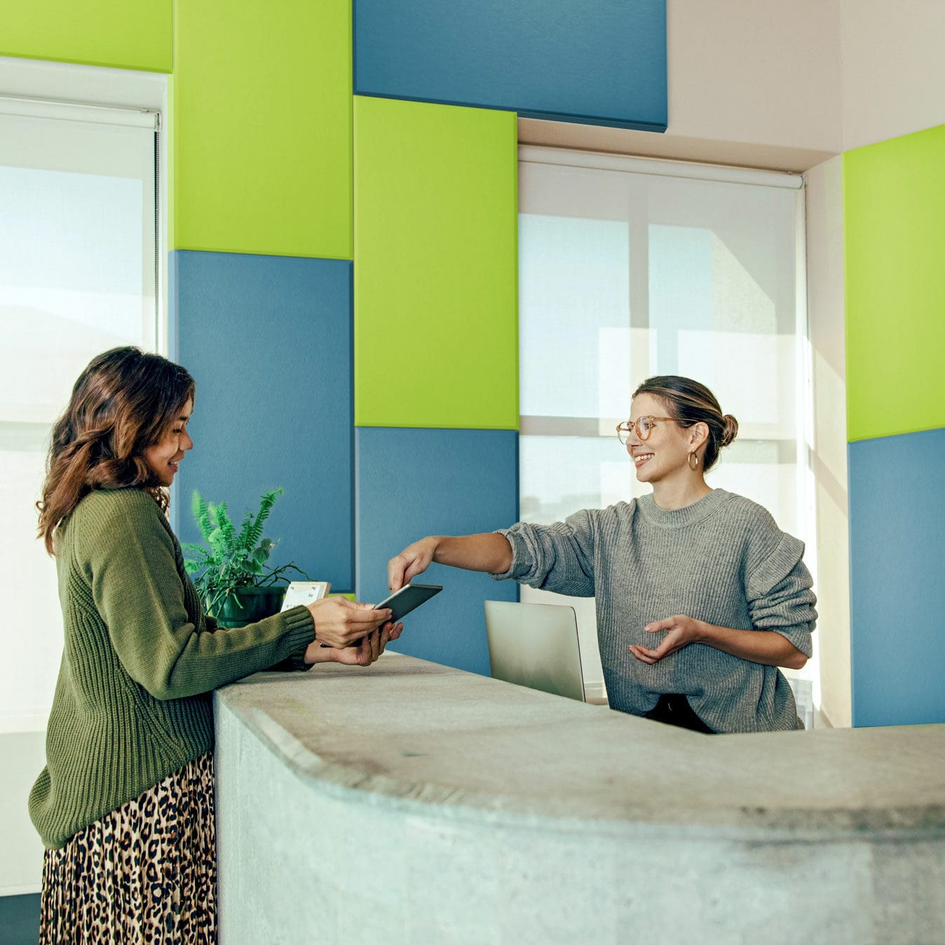 Two women interacting at store checkout counter with green and blue acoustic panels on wall.