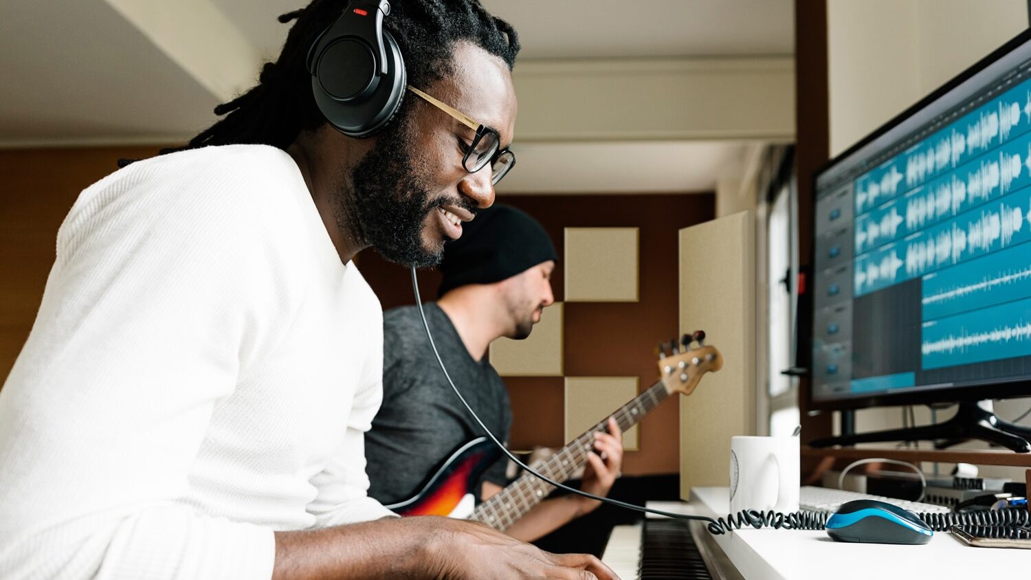 Two men playing instruments in a recording studio with beige acoustic panels on back wall.